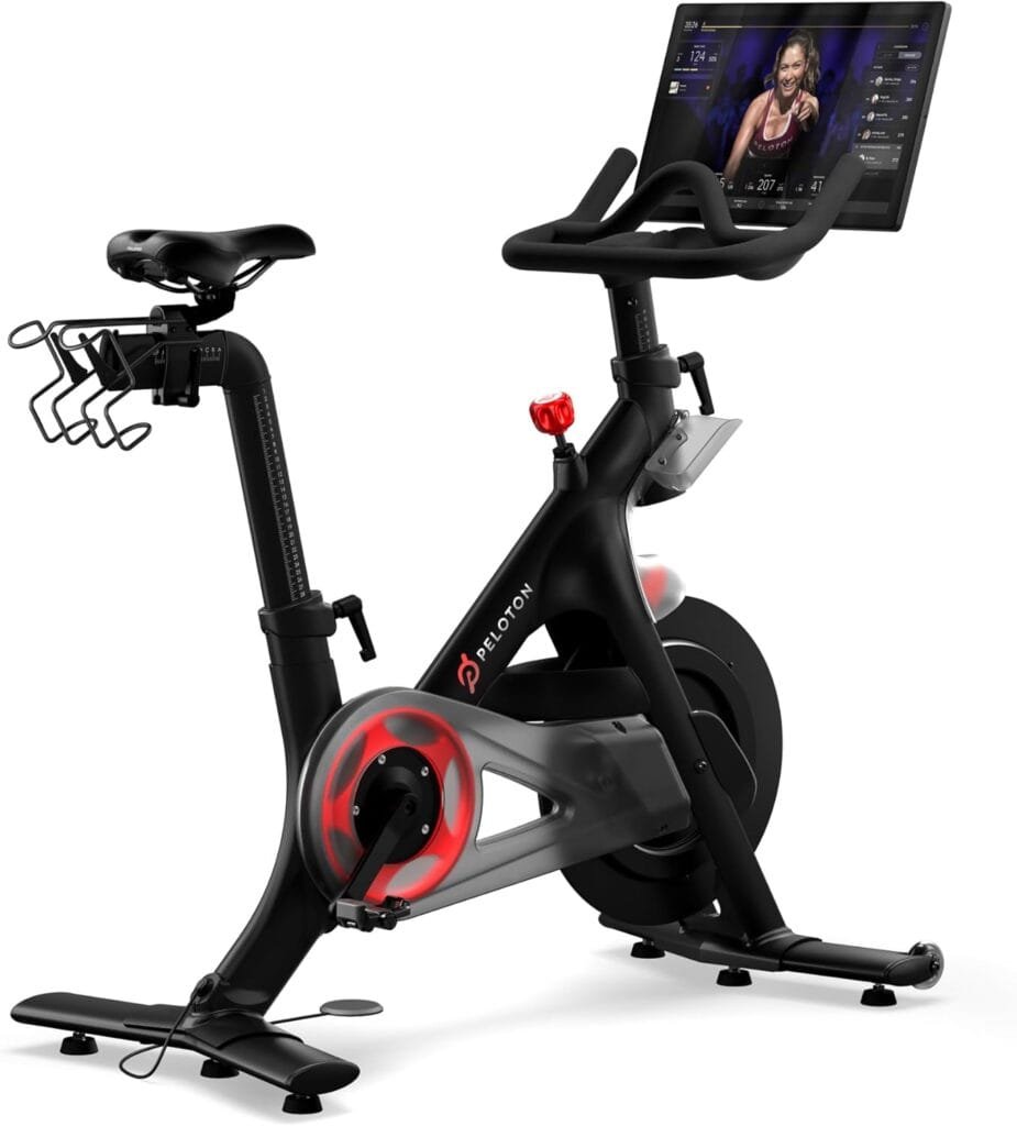 Peloton indoor exercise bikes: A stationary Peloton exercise bike with a mounted screen displaying a virtual instructor. The bike has a black frame, two water bottle holders, and red accents on the wheel.