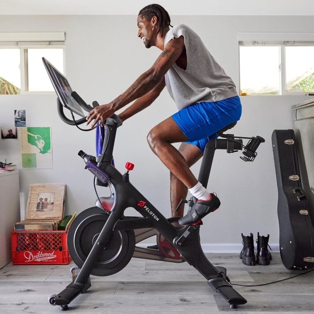 Peloton indoor exercise bikes: A person exercises on a Peloton stationary bike in a well-lit room with two small windows, a crate with books, a guitar case, and various items in the background.