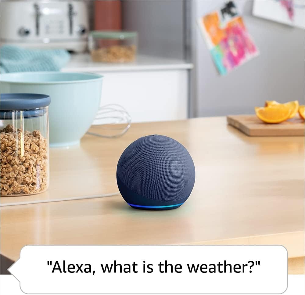 A Amazon Echo Dot 5th Gen smart speaker sits on a kitchen counter next to a cereal container and a mug, with a speech bubble asking, "Alexa, what is the weather?.