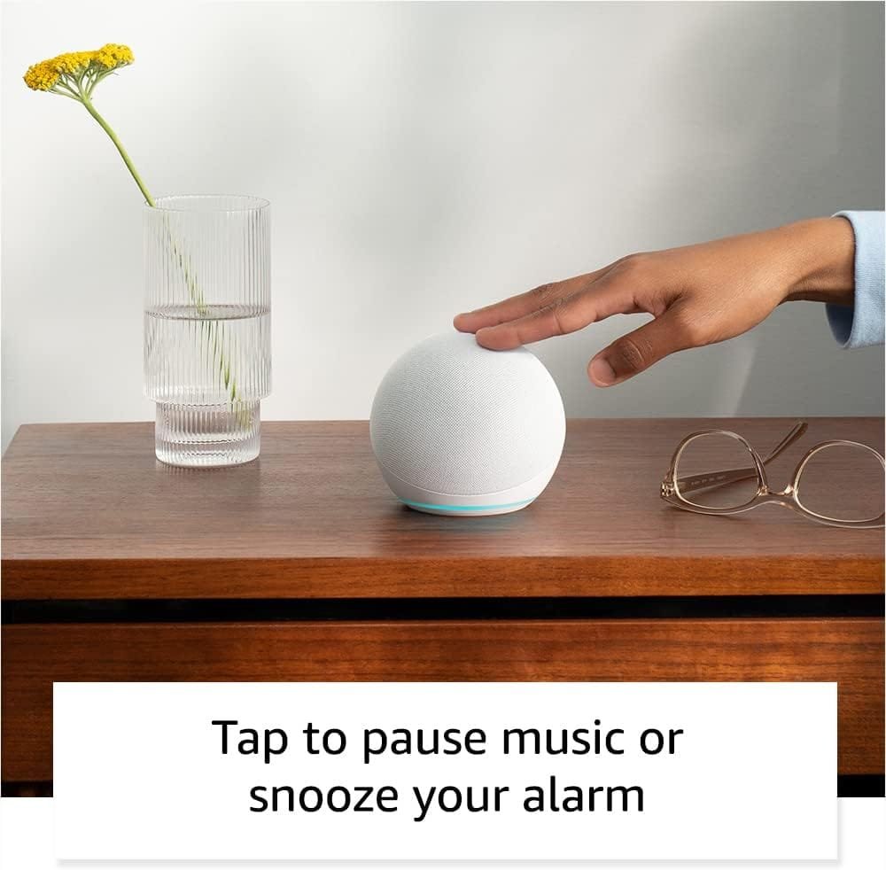 A hand taps a Amazon Echo Dot 5th Gen smart speaker on a wooden surface beside a glass of water and glasses, with text: "Tap to pause music or snooze your alarm.